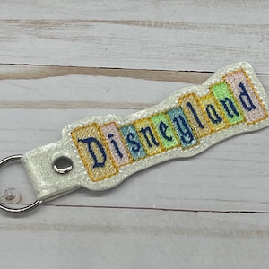 May include: White glitter keychain with a blue embroidered "Disneyland" banner. The banner is made of colorful fabric squares.