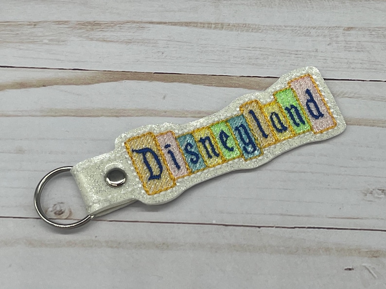 May include: White glitter keychain with a blue embroidered "Disneyland" banner. The banner is made of colorful fabric squares.