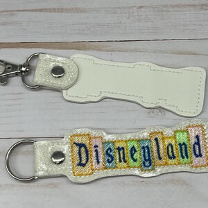 May include: White blank keychain with a silver key ring and a white keychain with the text "Disneyland" in colorful letters.
