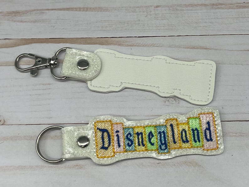 May include: White blank keychain with a silver key ring and a white keychain with the text "Disneyland" in colorful letters.