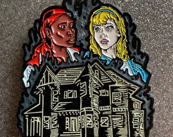 May include: A metal enamel pin featuring two figures above a dark house. On the left, a person with dark skin wears a red headscarf and shirt. On the right, a person with blonde hair wears a blue headband and dress. The house is black with gold lines.