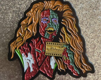 May include: A colorful enamel pin featuring a stylized, monstrous figure with long orange hair. The figure's face is split, with one side red and the other green. The figure holds a gold sign that reads "Metropol".