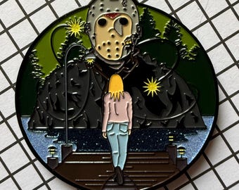 May include: Round enamel pin featuring a horror movie scene. The design includes a masked figure, a person walking away, a lake, and trees. The pin has a black, green, blue, and tan color scheme. The pin is a collectible item.