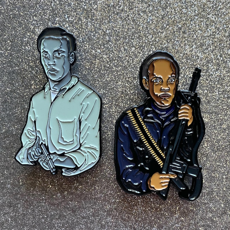 Op de afbeelding: Twee emaillepins die personages uit de film 'Do the Right Thing' voorstellen. De pin aan de linkerkant toont een lichtblauw shirt met het gezicht van een man. De pin aan de rechterkant toont een man in een donkerblauw jack die een geweer vasthoudt.