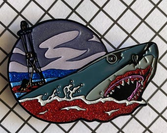 May include: Enamel pin featuring a shark with an open mouth, teeth bared, and a lighthouse in the background. The shark is teal, with a red and white mouth. The background is a dark blue sky with white clouds and a red glittery ocean.