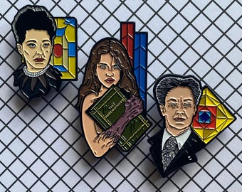 May include: Three enamel pins featuring characters from the movie The Three Mothers. The first pin is a woman with dark hair and a black dress. The second pin is a woman with long brown hair holding a book that says "The Three Mothers". The third pin is a man with dark hair wearing a suit and tie.