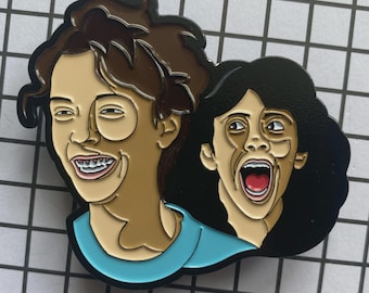 May include: Enamel pin featuring a cartoon illustration of two people with exaggerated expressions. One person is smiling widely, and the other is screaming with their mouth open. The pin has a blue, brown, and black color scheme.