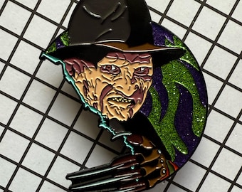 May include: Enamel pin featuring a stylized depiction of Freddy Krueger. The pin showcases the character's iconic hat, scarred face, and bladed glove, set against a green and purple glitter background. The pin is black, brown, red, green, purple, and tan.