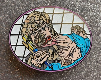 May include: A round enamel pin featuring a grotesque, stylized illustration of a figure with exposed flesh and a blue garment. The figure is holding a pen and a stack of yellow and blue pencils. The pin has a pink border and a white and black grid background.