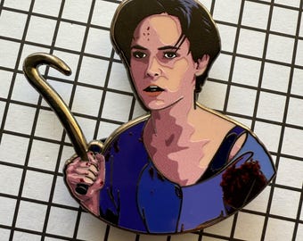 May include: Enamel pin featuring a stylized illustration of a person holding a golden hook. The person is depicted with dark hair and a blue top, with a pinkish skin tone. The pin has a gold border and is set against a black and white grid.