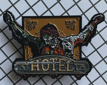 May include: A metal pin featuring a zombie with outstretched arms, set against a gold-colored background with the word "HOTEL" at the bottom. The zombie has exposed flesh and a decaying appearance, with a black and white grid background.