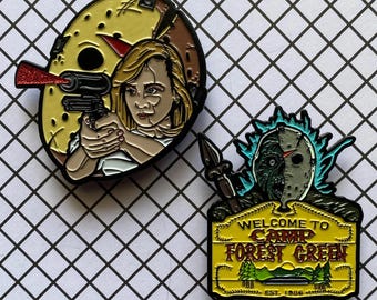 May include: Two enamel pins featuring a horror movie theme. The first pin is a yellow and red mask with a woman holding a gun. The second pin is a green and blue mask with a sign that reads "Welcome to Camp Forest Green Est. 1986".