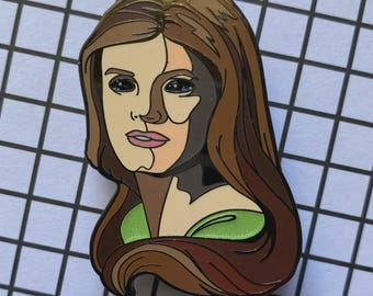 May include: Enamel pin featuring a stylized portrait of a woman with long brown hair and a green top. The face is divided into sections of light and dark colors, with gold accents on the hair and edges. The pin is set against a grid background.