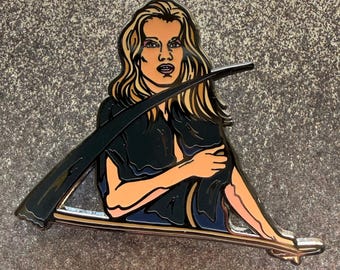 May include: Enamel pin featuring a seated figure with long blonde hair, wearing a black outfit, and holding a scythe. The pin has a gold outline and is set against a textured gray background.