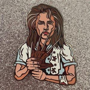May include: Enamel pin featuring a woman holding a brown glove with sharp metal claws. The woman has long brown hair and is wearing a white shirt with blue accents. The pin has a glittery silver background.
