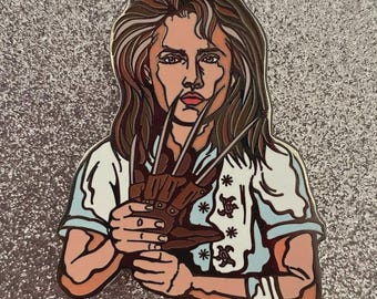 May include: Enamel pin featuring a woman holding a brown glove with sharp metal claws. The woman has long brown hair and is wearing a white shirt with blue accents. The pin has a glittery silver background.