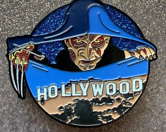 May include: A round enamel pin featuring a horror movie character with a blue hat and a scarred face. The pin has the word "HOLLYWOOD" in white letters and a starry background.