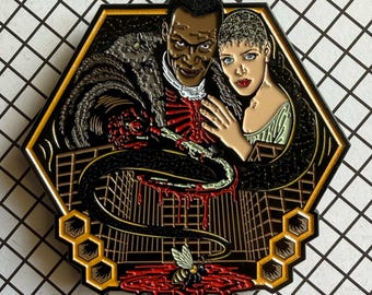 May include: A hexagonal enamel pin featuring a dark-skinned figure in a fur-trimmed coat and a light-skinned figure with short hair. A long, dark, blood-dripping tentacle extends downward toward a small bee resting in a pool of red liquid.