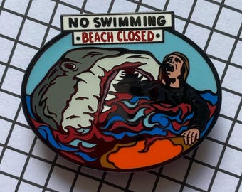 May include: A colorful enamel pin depicting a shark attacking a person in the water. The pin has a blue background with red and white waves. The text "NO SWIMMING - BEACH CLOSED -" is printed on a white banner above the shark.