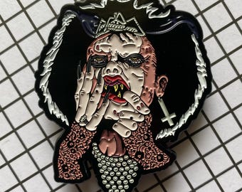 May include: A metal enamel pin featuring a stylized figure with pale skin, sharp yellow fangs, and long black fingernails. The figure wears a pearl necklace, a patterned garment, and an inverted cross earring, set against a black background.
