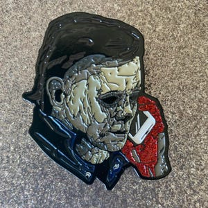 Può includere: Spilla smaltata raffigurante Michael Myers della serie di film horror Halloween. La spilla ha un contorno nero, con un volto argentato e grigio, capelli neri e una sezione rossa glitterata sul lato destro.