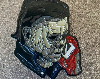 May include: Enamel pin featuring a stylized depiction of Michael Myers from the Halloween horror film series. The pin has a black outline, with a silver and gray face, black hair, and a red, glittery section on the right side.