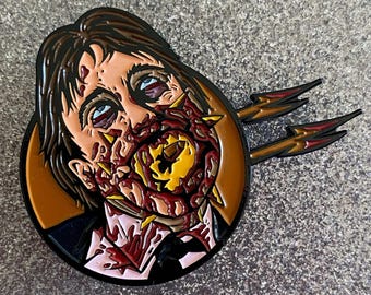 May include: A detailed enamel pin featuring a zombie with exposed flesh and a bloody, decaying face. The pin has a black outline, brown hair, and two lightning bolt accents. The zombie's face is a mix of red, yellow, and flesh tones.