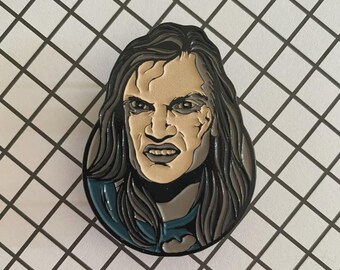 May include: A circular enamel pin featuring a detailed illustration of a person with long, dark, flowing hair and a stern facial expression. The figure has a pale, tan-colored face with defined features, dark eyes, and a blue garment visible at the bottom.