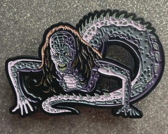 May include: A black, purple, and gray enamel pin featuring a creature with a reptilian body and a human-like face. The creature has long brown hair and is crawling. The pin has a detailed design and is likely a collectible item.