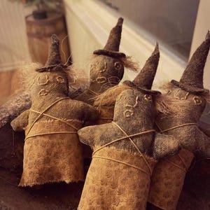 May include: A group of four handmade fabric dolls shaped like witches, each wearing a pointed hat and a tan skirt. The dolls are crafted from dark brown textured fabric with visible stitching for facial features and frayed edges on the skirts.