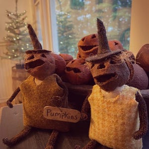 Puede incluir: Dos figuras de calabaza de tela hechas a mano con cabezas texturizadas de color marrón oscuro, sombreros puntiagudos y extremidades delgadas. Una figura lleva un chaleco amarillo mostaza y sostiene una etiqueta de madera "Pumpkins".