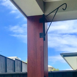 May include: A black metal plant hanger is attached to a wooden post. The hanger is curved and has a hook at the end. The post is painted a reddish brown color. The background is a blue sky with white clouds.