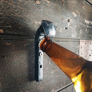 May include: A black metal bottle opener mounted on a wall with a brown beer bottle being opened.