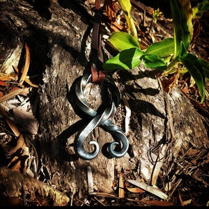 May include: A silver metal pendant in the shape of a stylized knot, hanging from a brown leather cord. The pendant is resting on a piece of weathered wood.