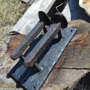 May include: Hand-forged iron door pulls with a rustic, hammered texture. The pulls feature a rectangular backplate with two protruding handles, each with a circular head. The dark metal contrasts with the natural wood surface.
