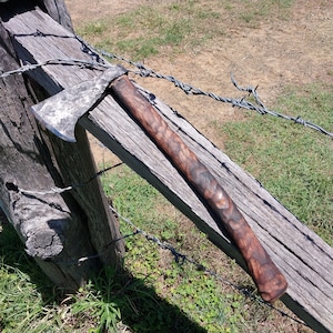 May include: A hand-forged axe with a wooden handle. The axe head is made of metal and has a rough, textured surface. The handle is made of wood and has a dark brown finish. The axe is resting on a wooden fence post.