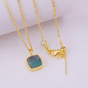 May include: A gold-colored necklace with a square, blue-green gemstone pendant. The pendant is set in a gold bezel. The necklace chain is delicate and has a lobster clasp closure. The necklace is displayed on a white surface.