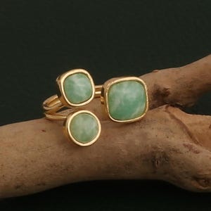 May include: A gold-toned ring with three light green, square and round gemstones. The ring is displayed on a wooden branch against a dark green background. The gemstones have a marbled appearance.