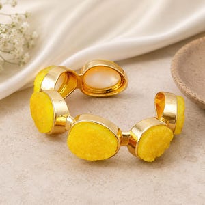 May include: A gold-coloured cuff bracelet with bright yellow, textured stones and a central, oval-shaped, off-white stone. The bracelet has a hinged design and is displayed on a neutral surface.