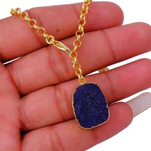 Puede incluir: Un collar de cadena dorada con un colgante azul oscuro de forma irregular. El colgante tiene un borde dorado. El collar se muestra sobre un fondo blanco.