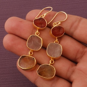 May include: Gold-toned dangle earrings featuring three natural gemstones in shades of red, pink, and yellow. The earrings have a hook closure and are held in a hand against a purple background. The gemstones are irregularly shaped.