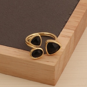 Może przedstawiać: Złoty pierścionek z trzema czarnymi kamieniami: owalnym i trójkątnym po jednej stronie oraz trójkątnym po drugiej. Pierścionek jest prezentowany na drewnianej powierzchni.