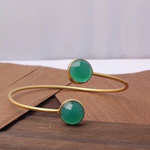 May include: A gold-toned cuff bracelet featuring two round, faceted, green gemstones. The bracelet has a simple, open design with the gemstones set at each end. The gemstones are a vibrant green color, and the metal has a polished finish.