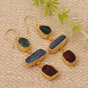 May include: Gold-tone earrings with a drop design. Each earring features three gemstones: a green oval, a blue rectangular, and a maroon oval. The gemstones are set in gold-colored bezels. The earrings are displayed on a beige surface.