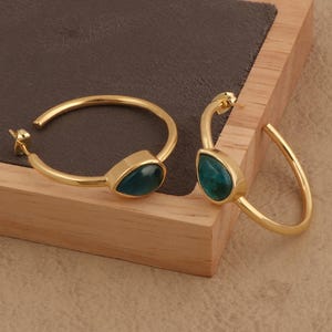 May include: A pair of gold-colored hoop earrings. Each earring features a teardrop-shaped, teal-colored stone set in a gold bezel. The earrings are displayed on a wooden surface, showcasing their design and color.