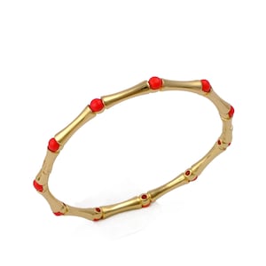 May include: A gold-toned bangle bracelet featuring a segmented bamboo-inspired design. The bracelet is accented with small, round, bright red beads placed at each joint between the smooth, metallic gold segments of the circular jewellery piece.