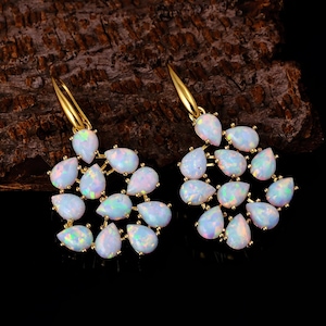 May include: A pair of gold earrings with clusters of pear-shaped, iridescent white opals. The opals are arranged in a floral pattern, with each earring showcasing a delicate design. The background is a dark, textured surface.