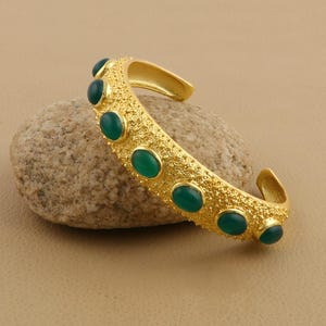 Könnte beinhalten: Ein goldfarbenes Armband mit einer texturierten, körnigen Oberfläche, das mit einer Reihe von sieben glatten, ovalen grünen Cabochon-Steinen besetzt ist, die entlang der äußeren Kurve des offenen Metallbandes angeordnet sind.