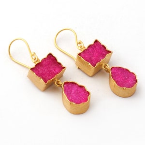 May include: A pair of gold-colored earrings with a square top and a teardrop-shaped bottom. The earrings feature a vibrant pink, textured stone set within the gold frame. The earrings hang from a simple hook.