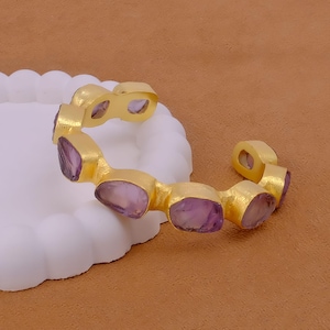 May include: A gold-coloured cuff bracelet with irregularly shaped, light purple gemstones. The bracelet has an open design and is resting on a white, cloud-shaped surface. The background is a solid, warm brown.